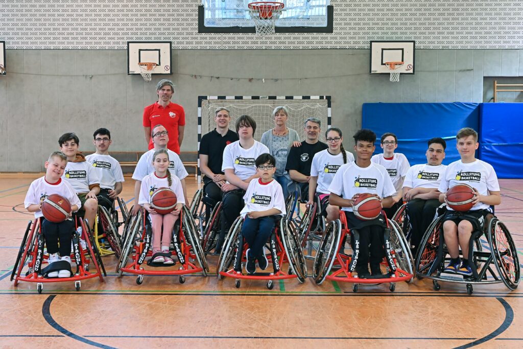 14 Rollstuhlbasketball-Talente sitzen unter einem Basketball-Korb für ein Teamfoto zusammen. Vier Mädchen und Jungen halten Basketballbälle in der Hand.