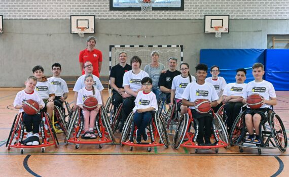 14 Rollstuhlbasketball-Talente sitzen unter einem Basketball-Korb für ein Teamfoto zusammen. Vier Mädchen und Jungen halten Basketballbälle in der Hand.