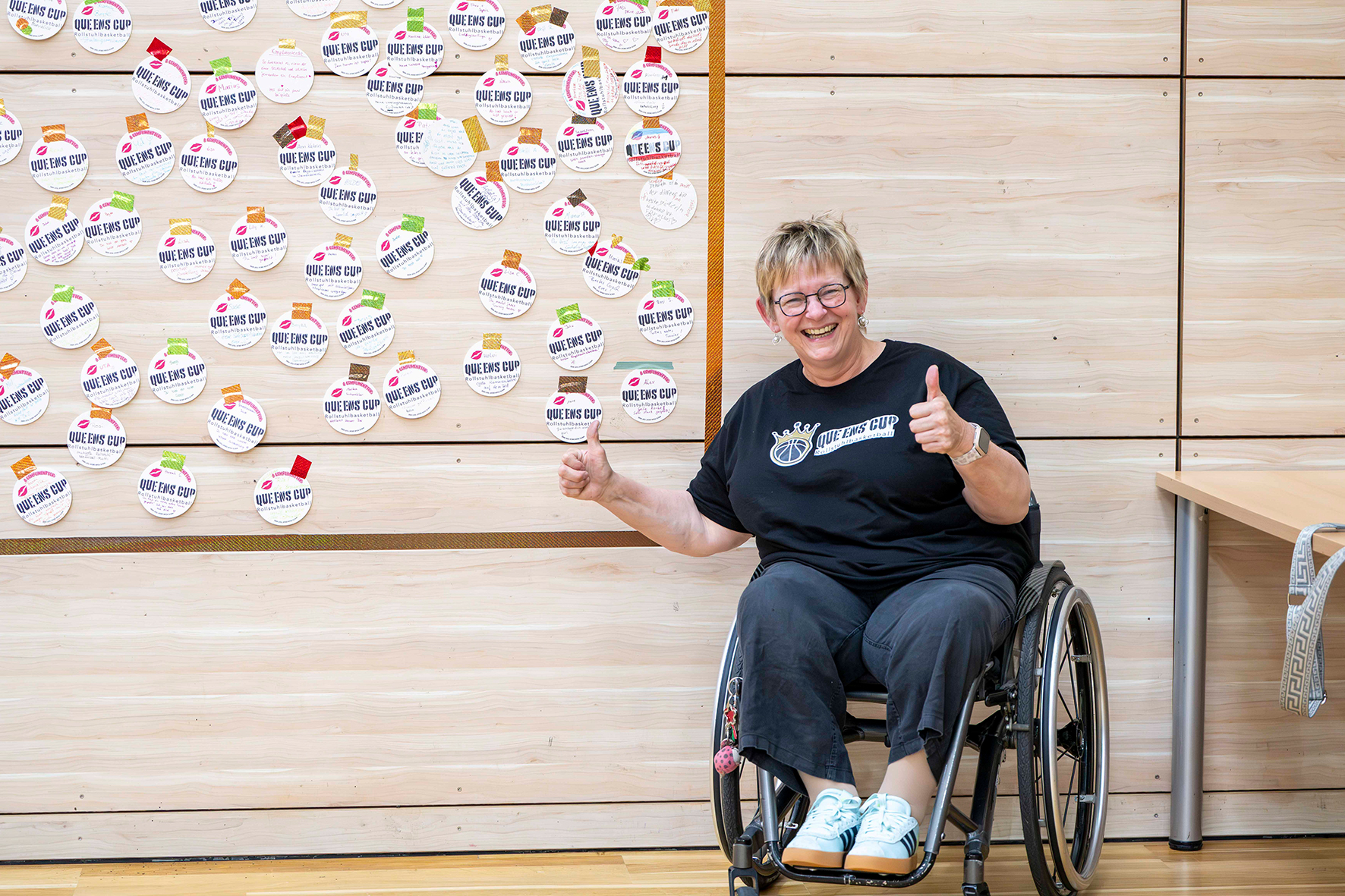 Eine Frau im Rollstuhl sitzt vor einer Pinnwand mit Stickern, auf denen Komplimente geschrieben sind. | Foto: Volker Ballisch