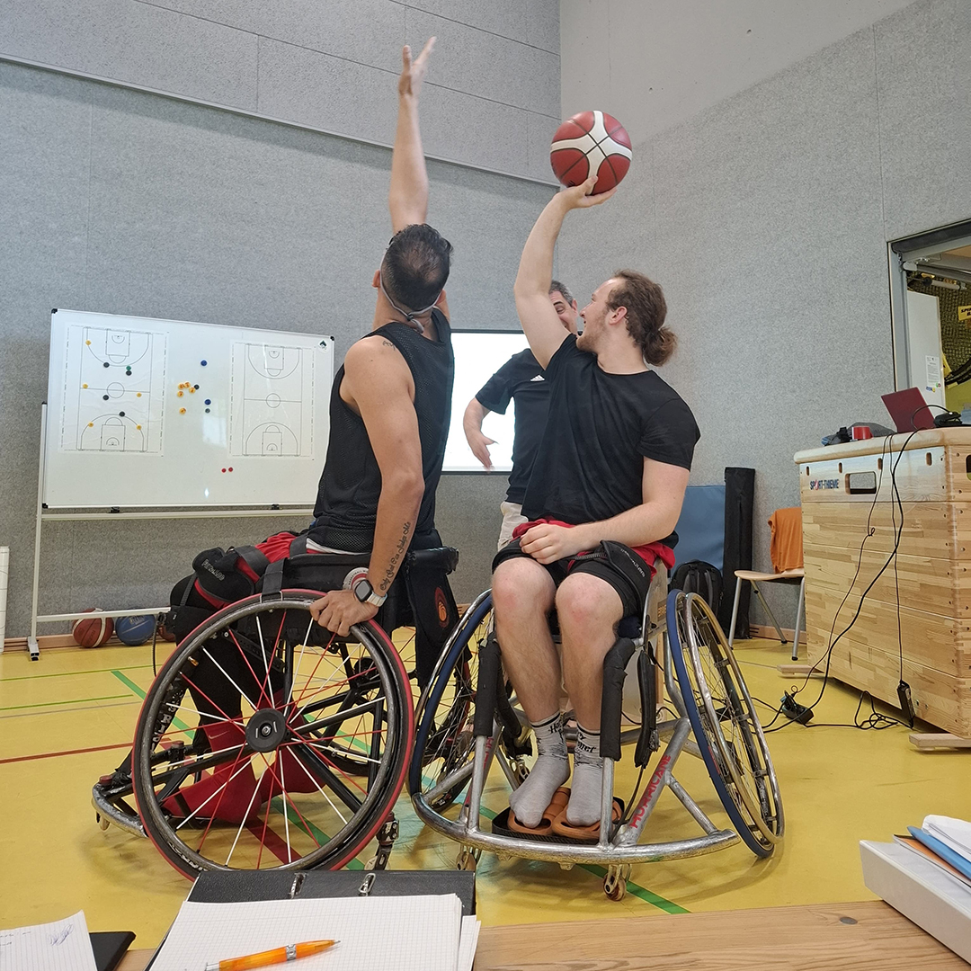 Zwei Rollstuhlfahrer beim Trainerlehrgang, von denen einer einer Basketball hochhält